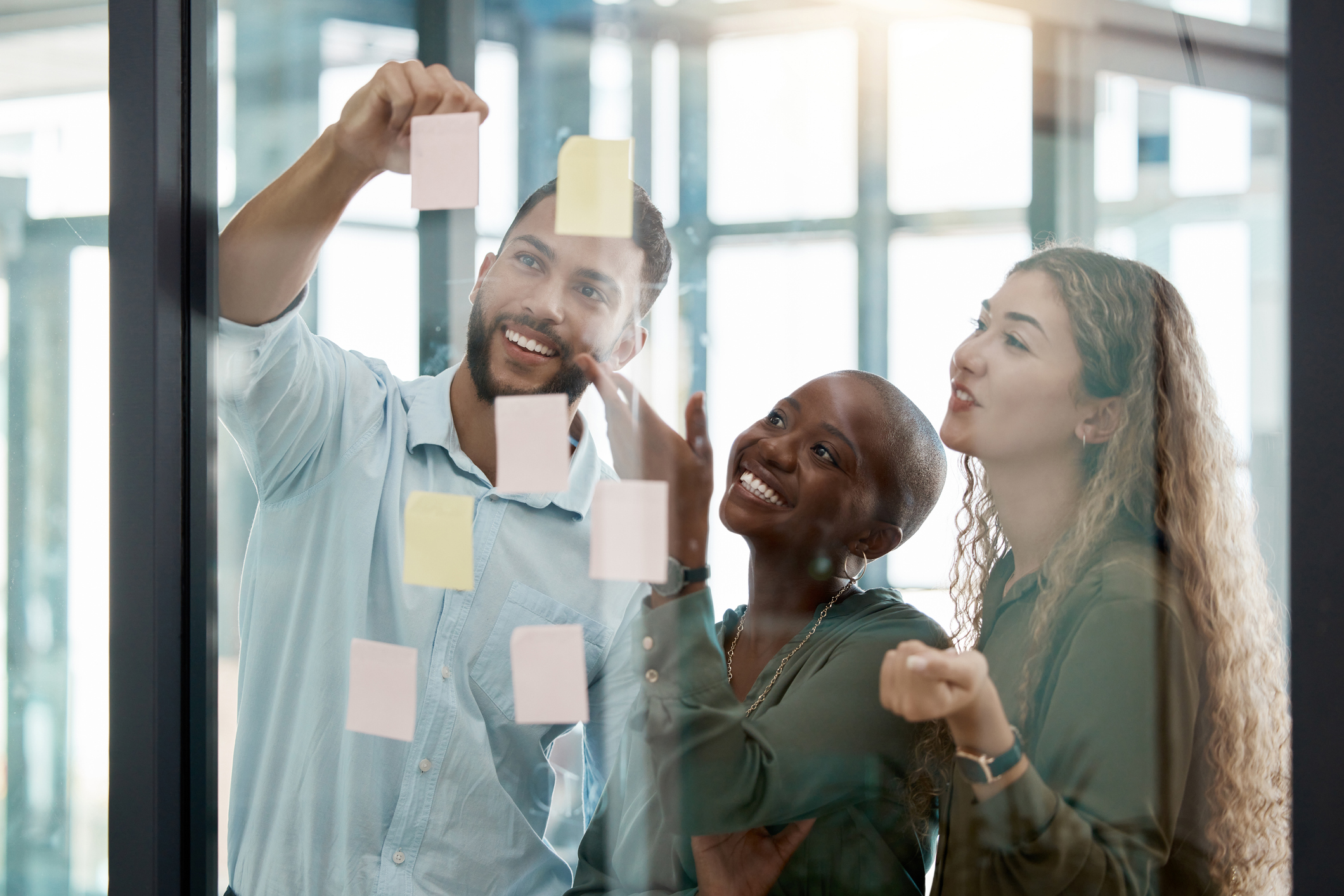 Photo of a diverse team in a business brainstorming strategy for planning and sticky notes. Three people sharing ideas, planning and scheduling work tasks in teamwork and delegation at office.