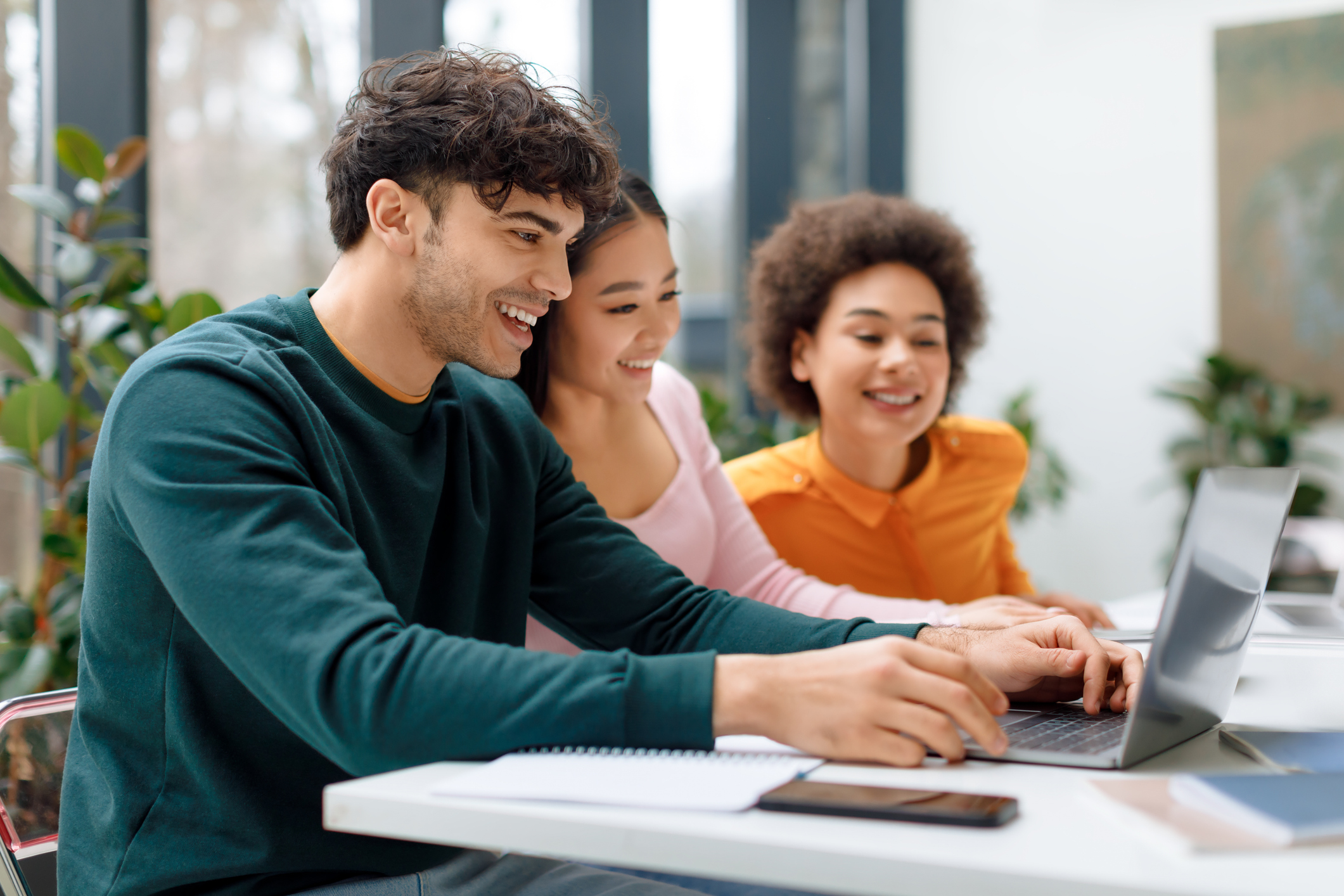 Three joyful students from different ethnic backgrounds engage in collaborative study session using laptop, surrounded by lush indoor plants