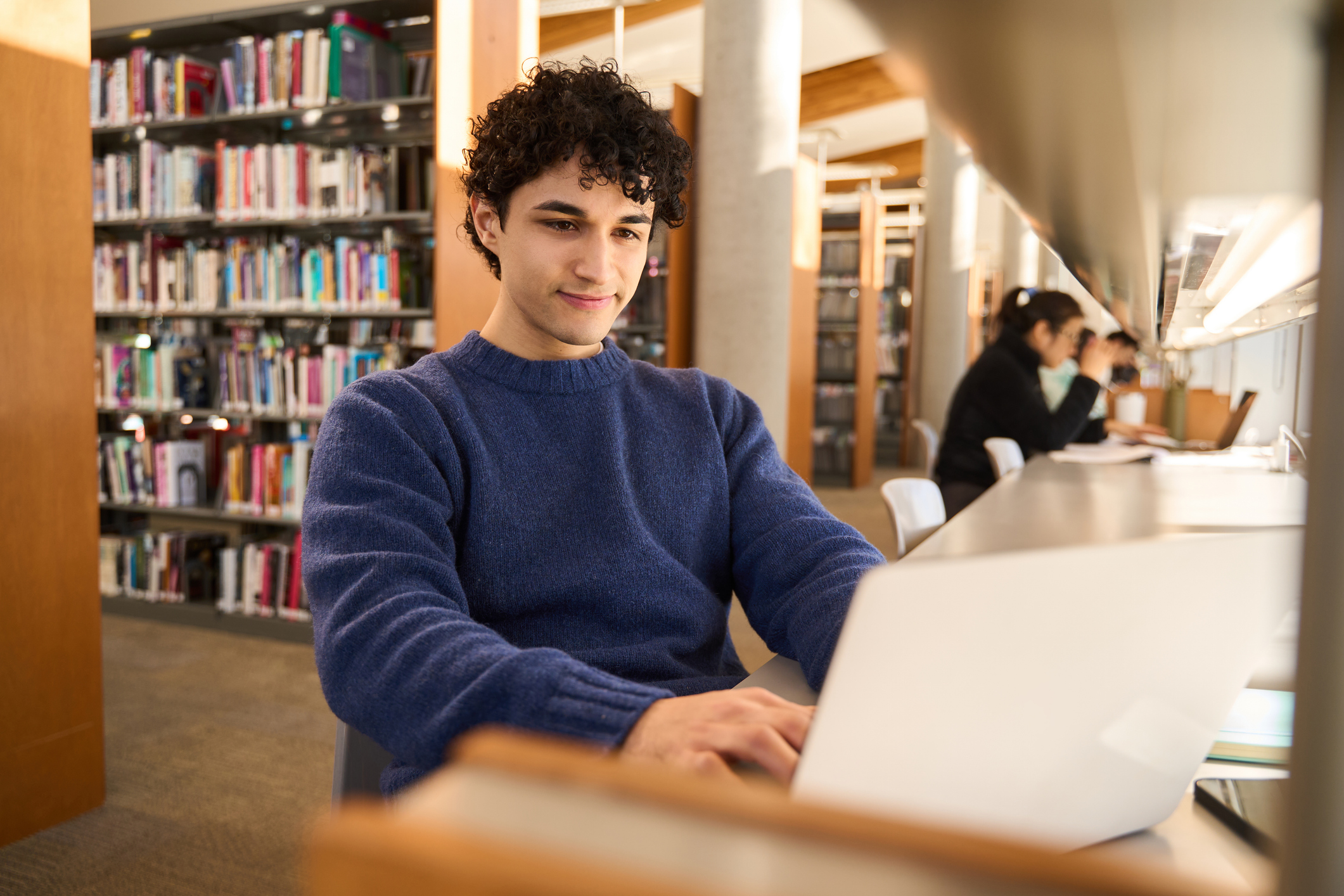 Photo of Young man smiling while working on laptop in public library with other students studying in background