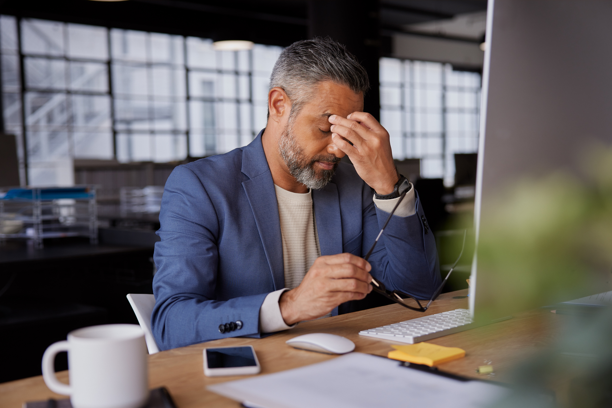 Photo of businessman holding his head in stress, sitting at a desk with computer and documents