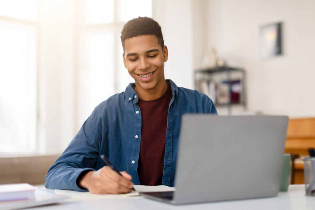 Content young male smiling as he writes notes with a pen from his laptop, sitting at a well-lit desk